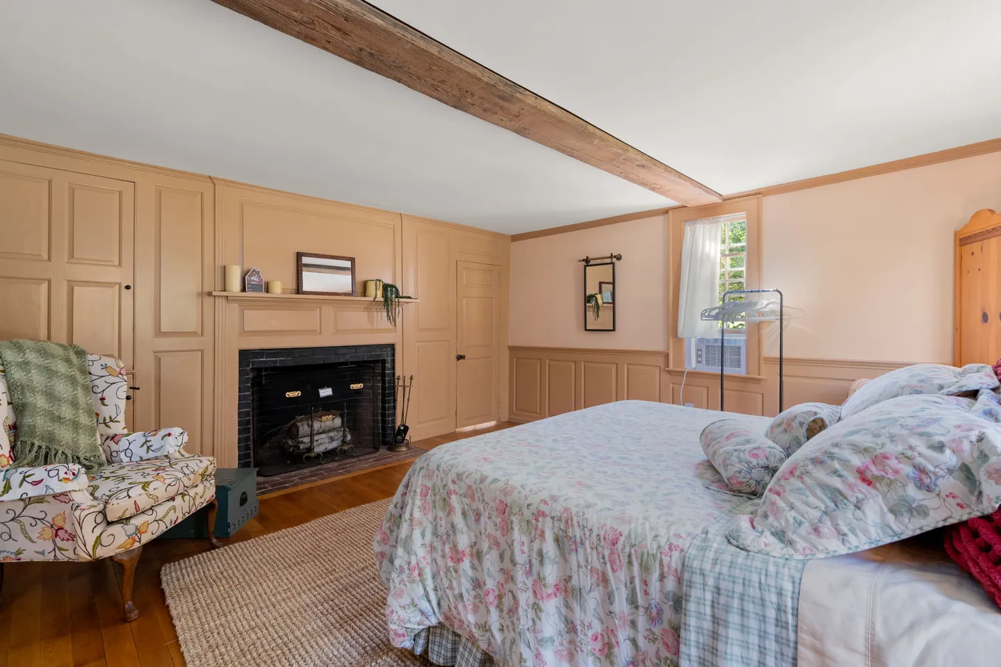 Bedroom with an exposed wooden ceiling beam, a fireplace surrounded by paneled walls, a floral patterned armchair, and a made bed.