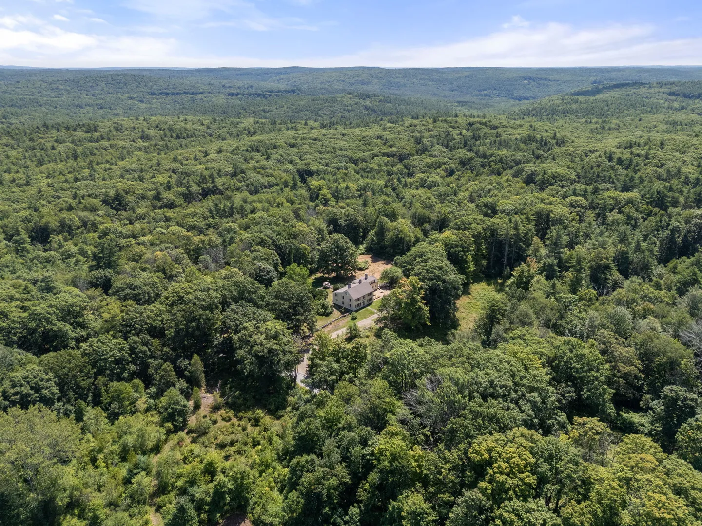 Aerial view of a house nestled in a dense green forest surrounded by rolling hills under a blue sky.