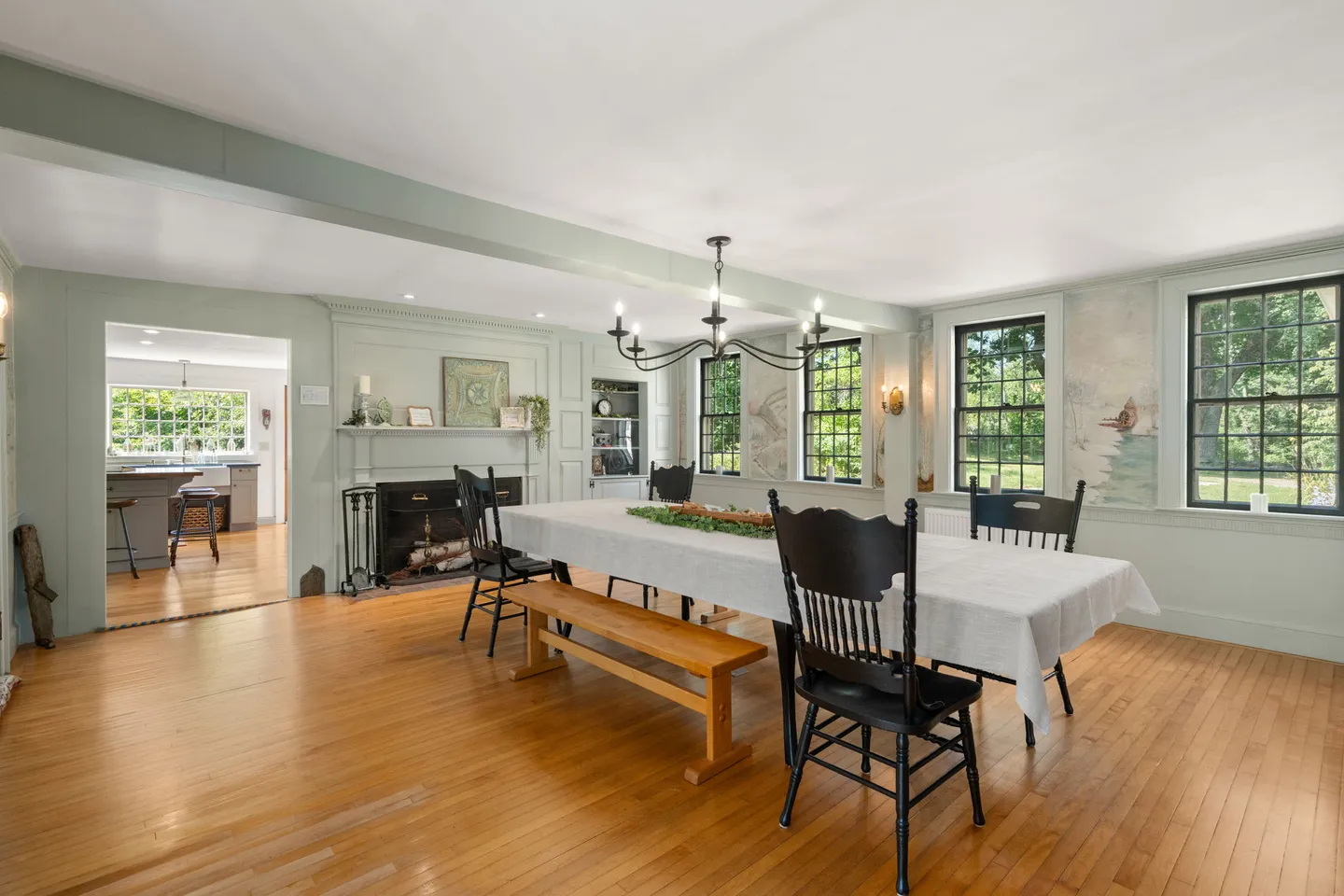 Spacious dining room featuring a long table with a white tablecloth, wooden bench, black spindle chairs, a fireplace, and views into an adjacent kitchen.
