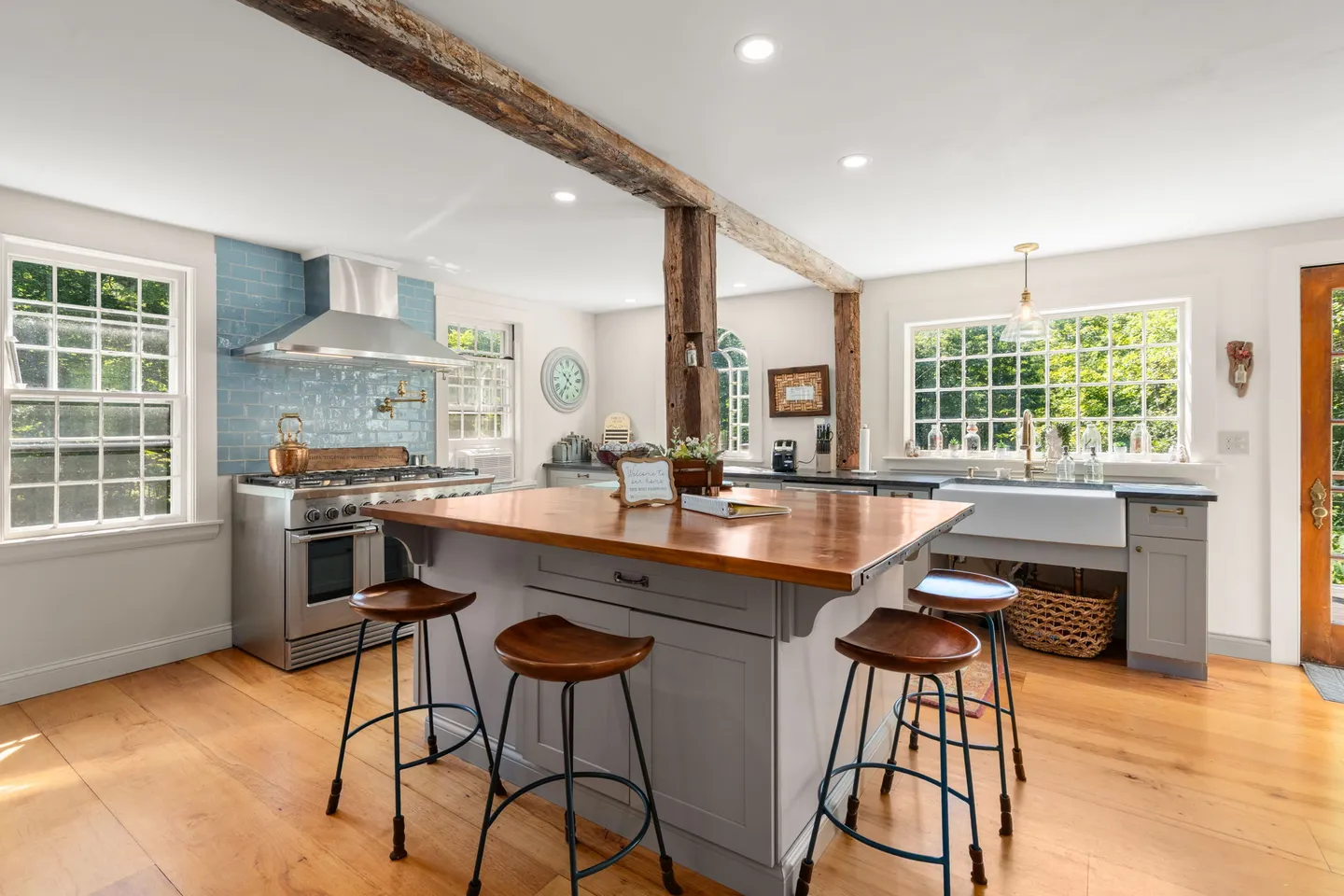 Bright kitchen featuring a wood-topped island with barstools, exposed rustic wooden ceiling beams, light wood floors, and multiple windows.