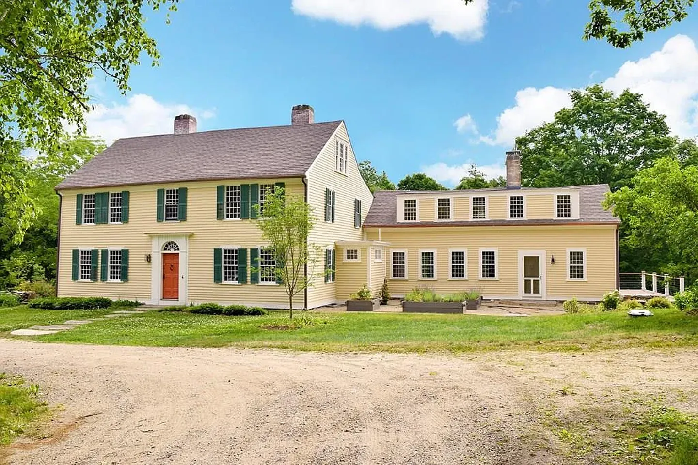 Exterior front view of a two-story yellow colonial-style home with green shutters, a reddish-orange front door, and a connected wing set against a blue sky.