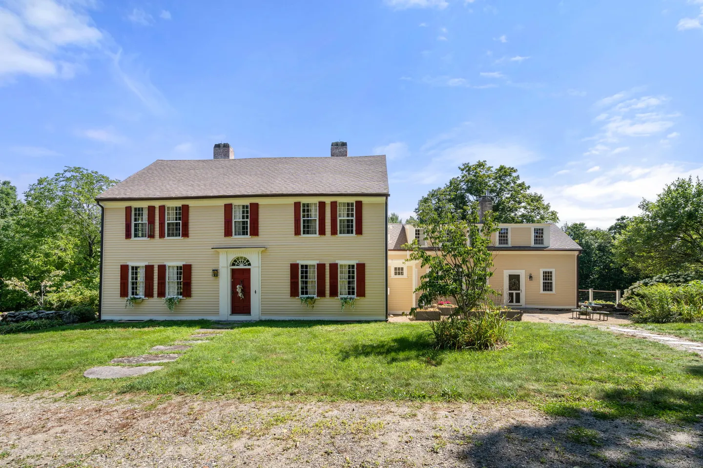 Exterior front view of a two-story yellow house with red shutters and an attached side wing on a sunny day.