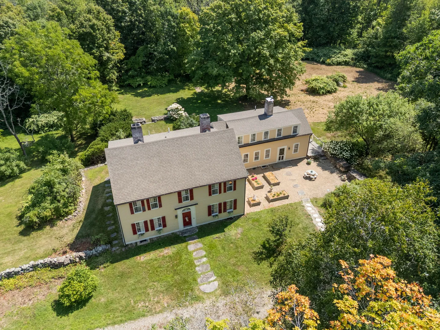 Aerial view of a two-story yellow historic home with red shutters, stone chimneys, and a stone patio surrounded by lush green trees and lawn.