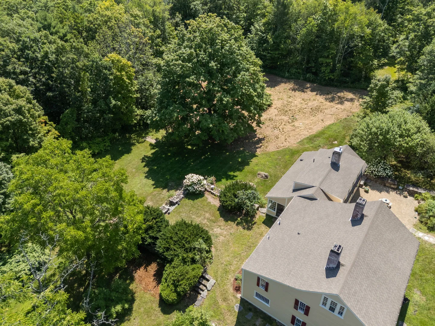 Aerial view of a multi-story home with shingled roofs and chimneys surrounded by dense green woods and lawn.