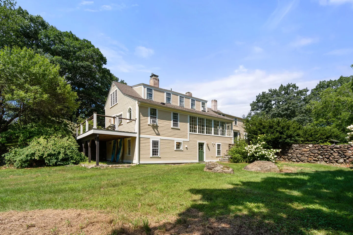 Exterior rear view of a two-story light yellow clapboard home with an elevated deck, stone wall, and spacious green lawn.