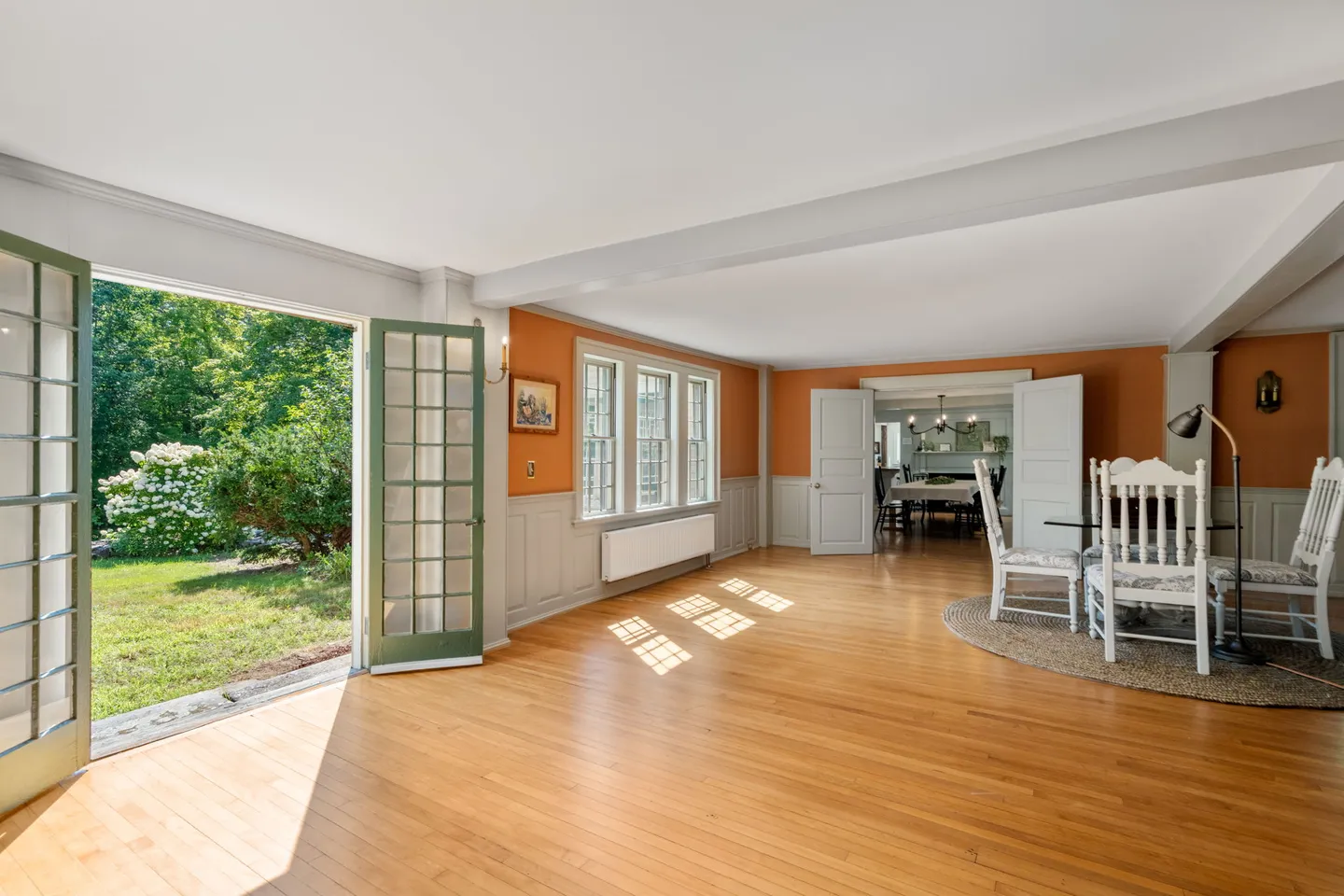 Sunlit room with light wood floors, terracotta walls, wainscoting, and French doors opened to a lush green yard.