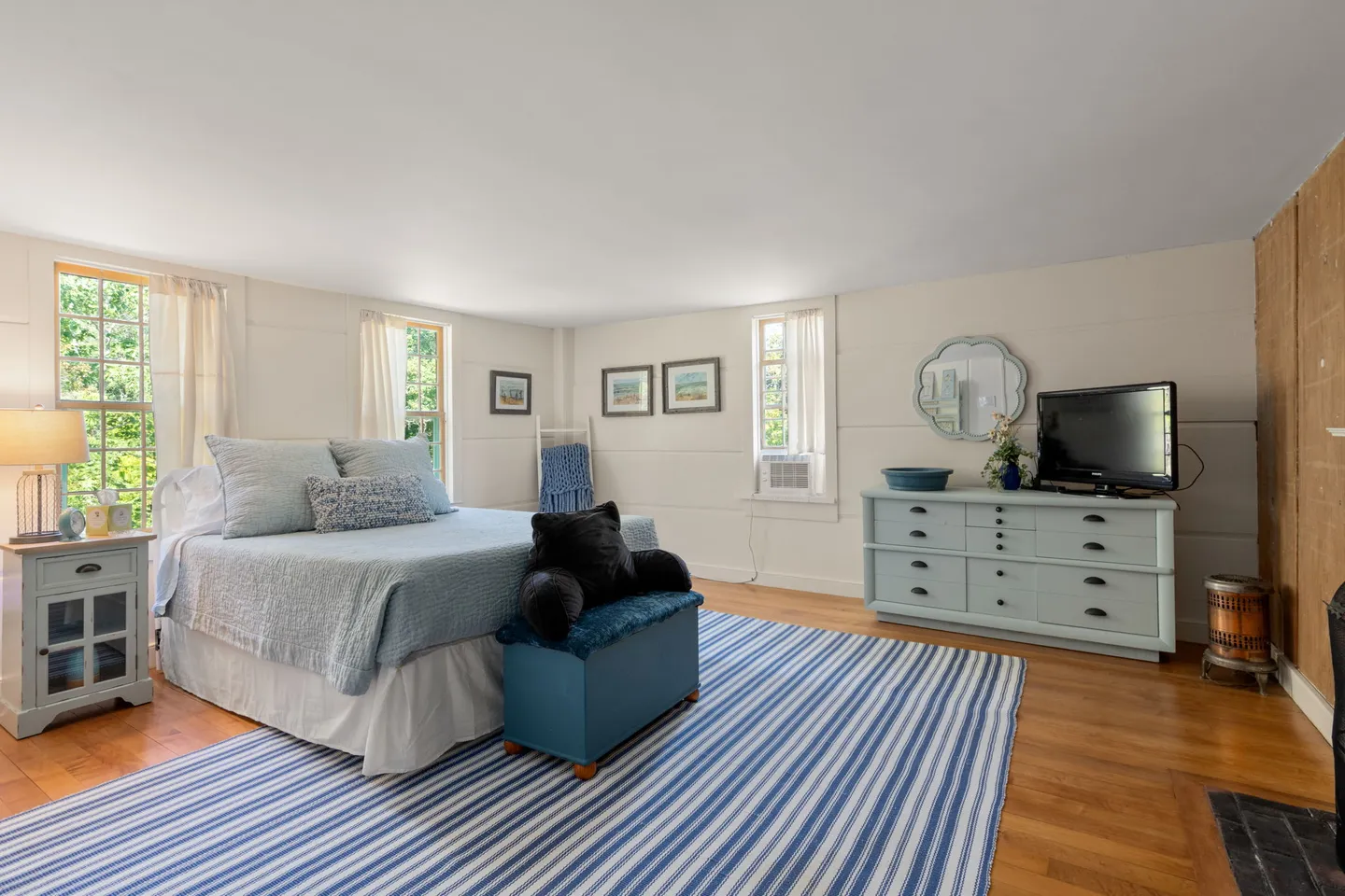 Bedroom with hardwood floors, a large blue and white striped rug, a bed dressed in light blue bedding, a nightstand, and a light blue dresser with a television.