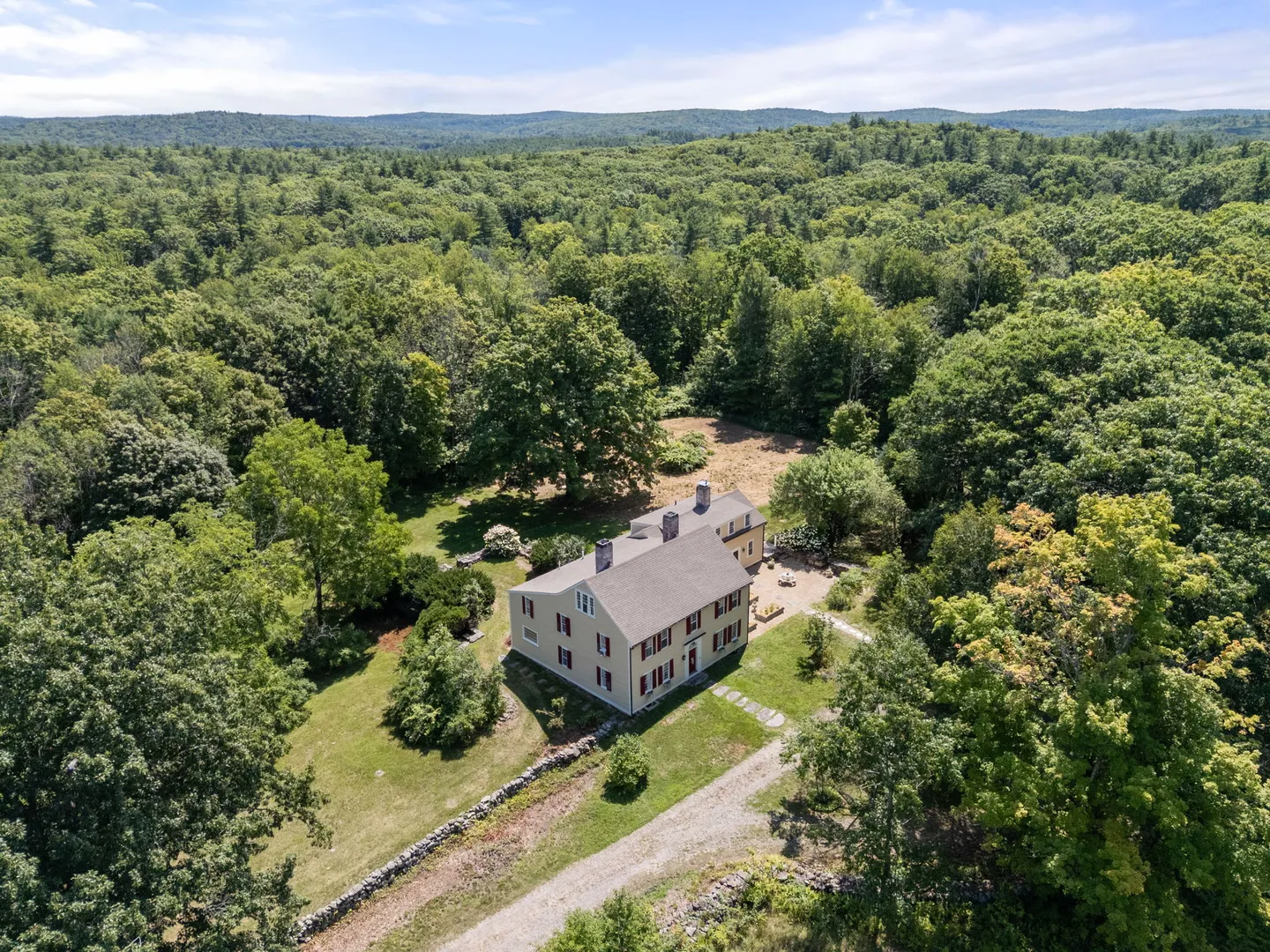 Aerial view of a multi-story home surrounded by dense forest, with a dirt driveway and stone walls.