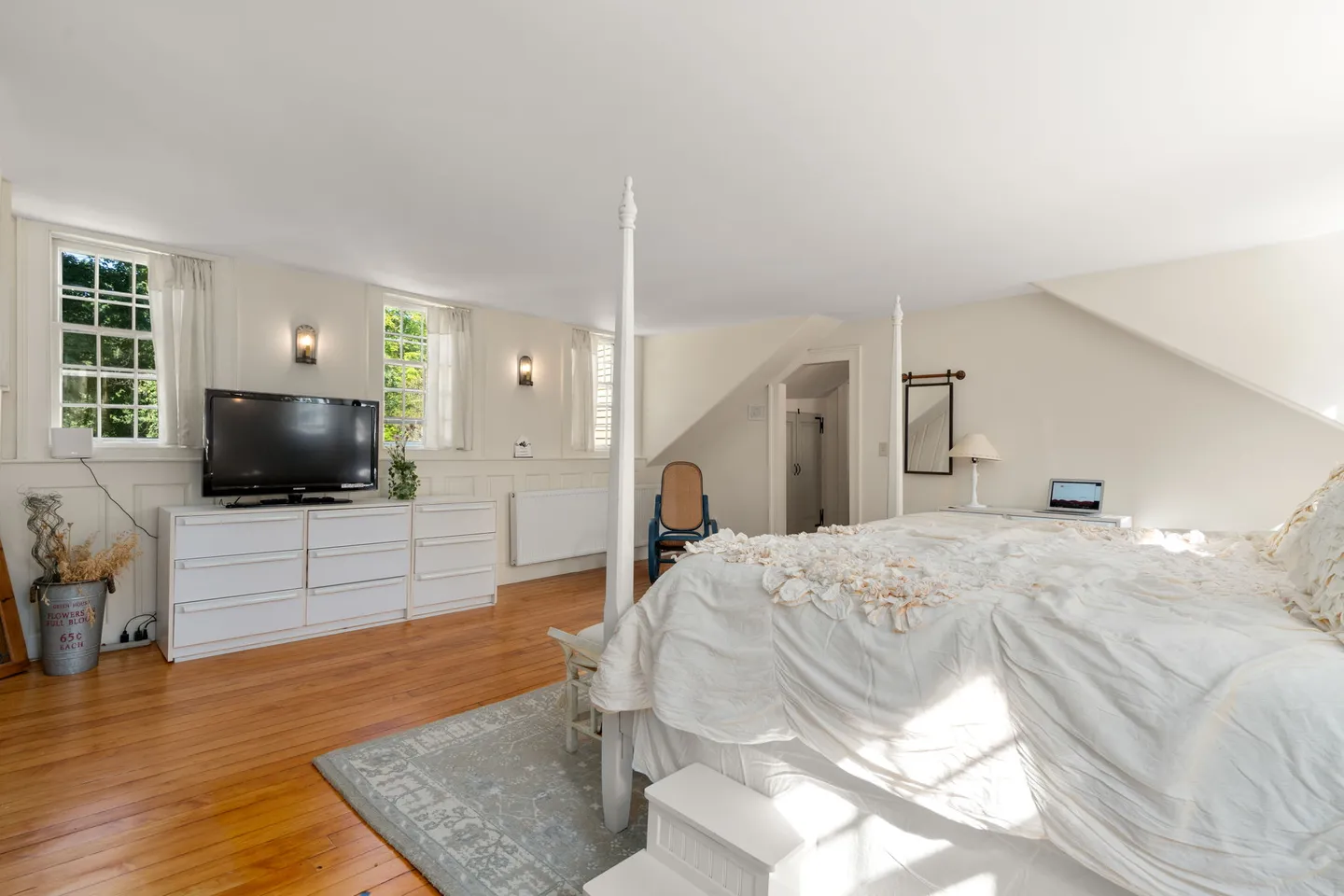 Bedroom with wooden floors, white four-poster bed, white dressers with a TV, and windows along a pale wall.