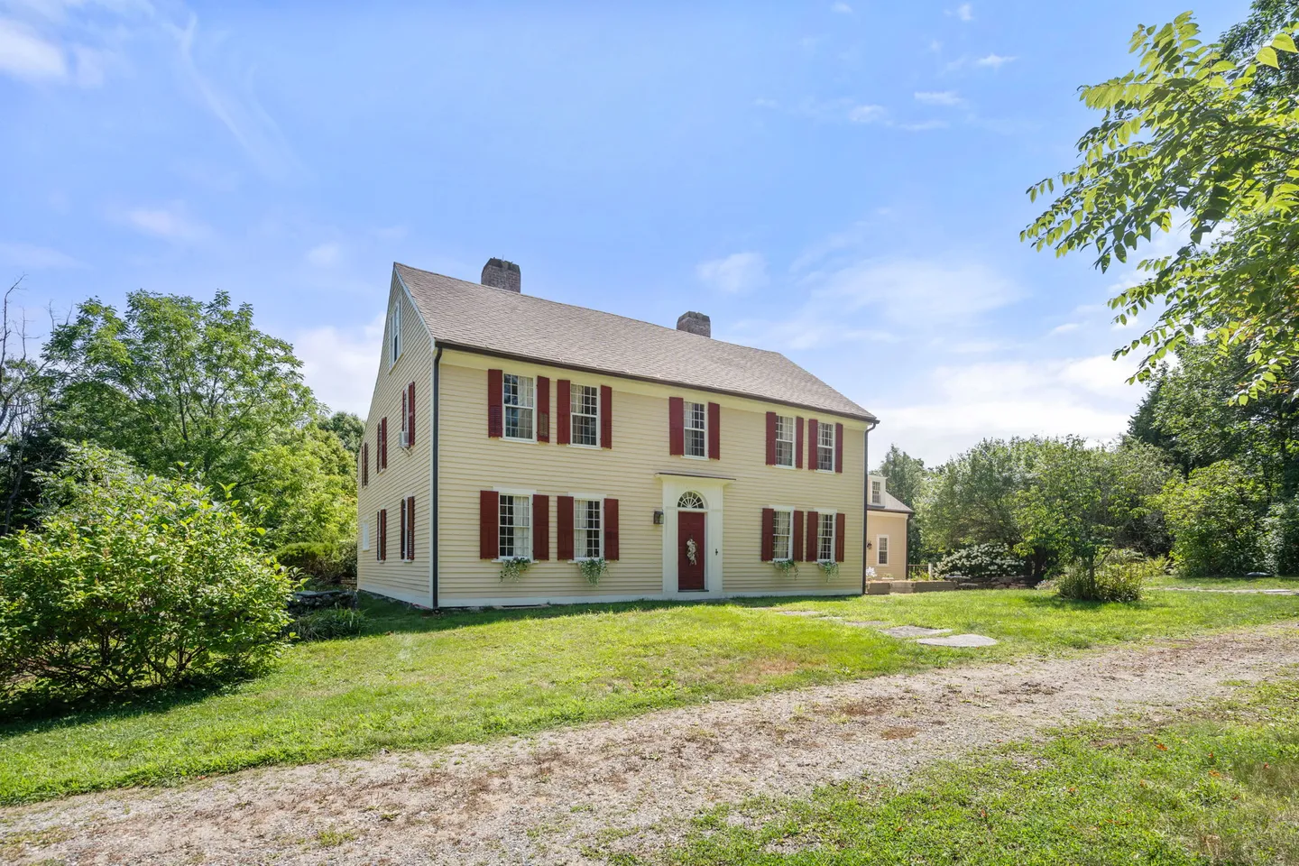 Exterior front view of a two-story yellow colonial-style home with red shutters and a gabled roof set on a grassy yard.