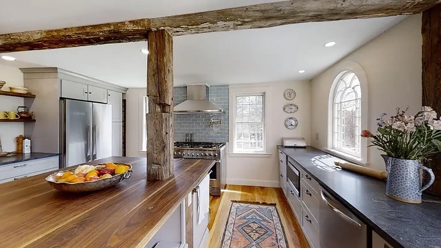 Kitchen interior featuring a rustic wooden support beam, wood-topped island, stainless steel appliances, and an arched window.