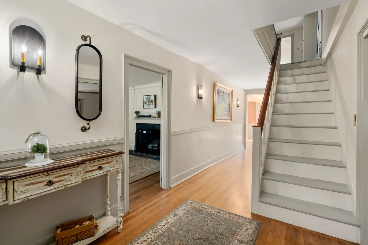 Hallway foyer with wood flooring, light painted stairs with a wooden handrail, a console table with an oval mirror, and a doorway leading into an adjoining room.