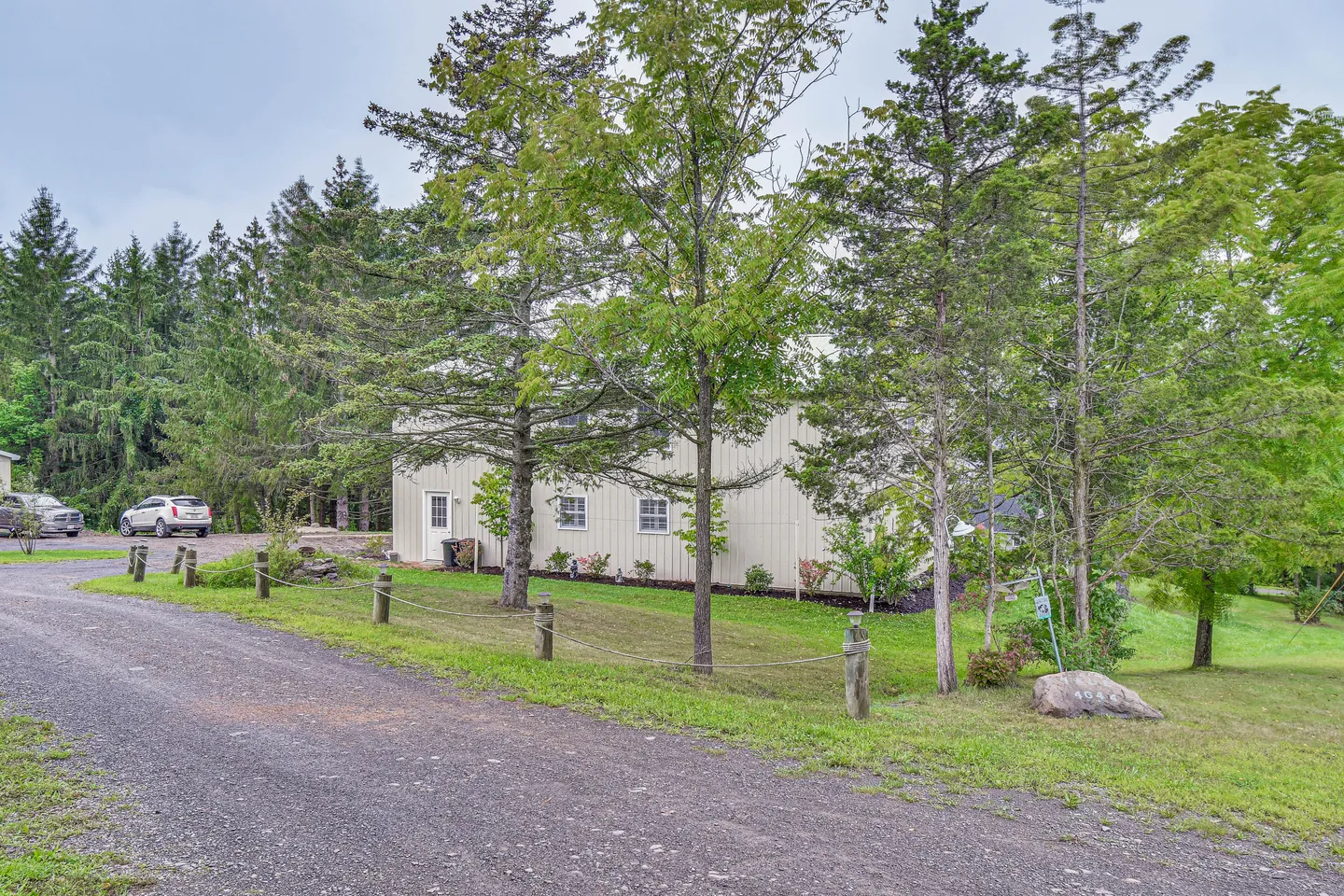 Exterior view of a light-colored metal building with small windows and a door, set behind a gravel driveway, grassy lawn, and trees.