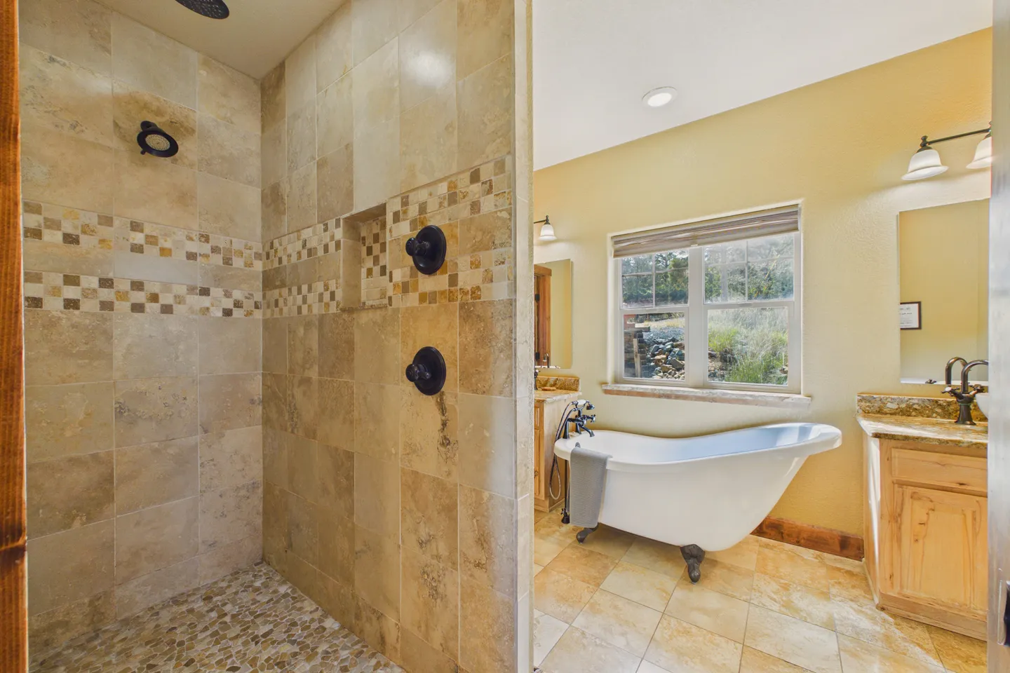 Bathroom showing a walk-in tile shower with pebble floor on the left, a clawfoot soaking tub beneath a window, and vanity cabinets.