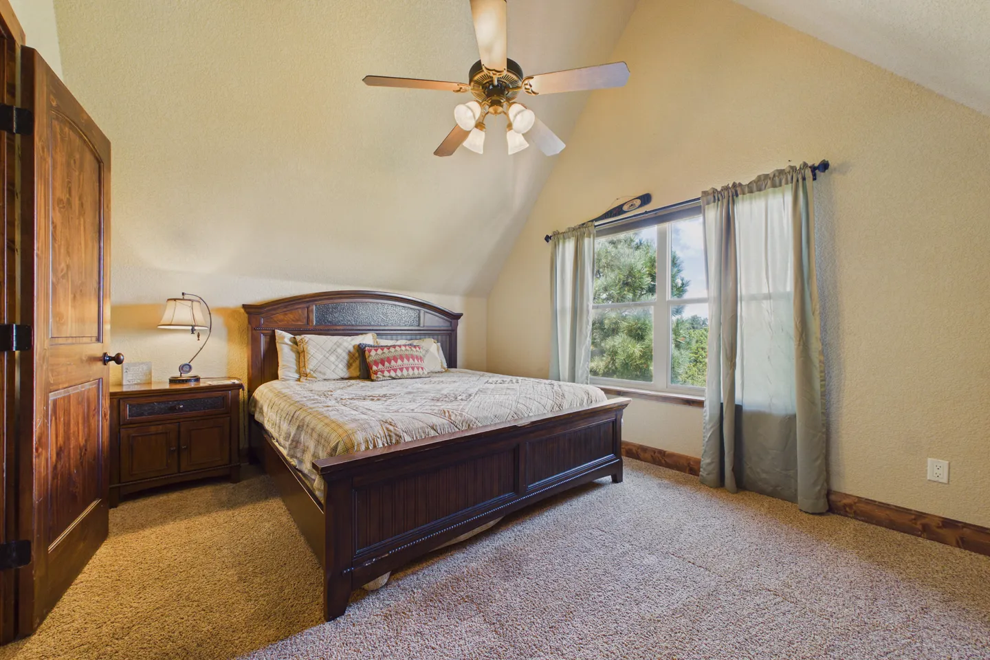 Bedroom with vaulted ceiling, wooden bed frame, ceiling fan, carpet, and a window with green curtains.