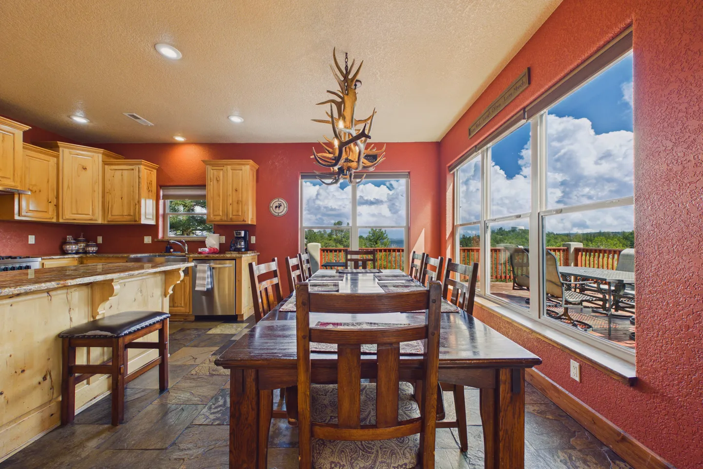 Dining area with a large wooden table, antler chandelier, and adjacent open kitchen with light wood cabinetry.