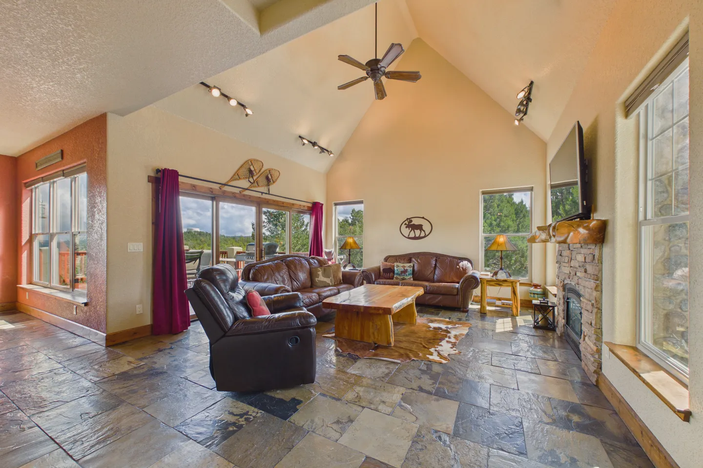 Living room with vaulted ceiling, stone-look tile flooring, leather seating, a stone fireplace, and large sliding glass doors.