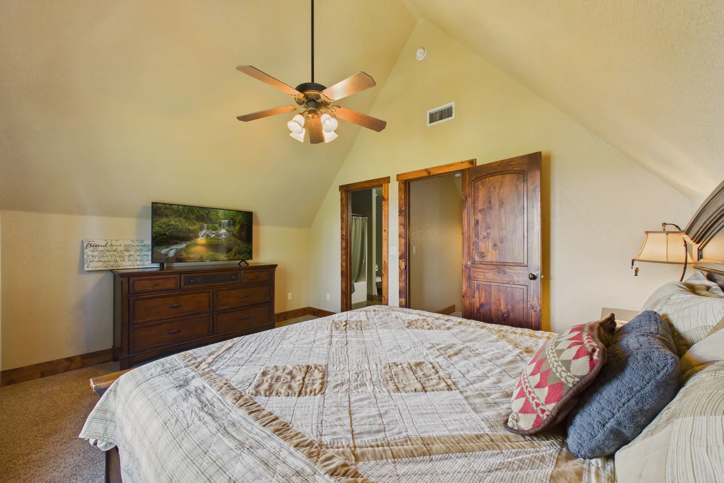 Bedroom with vaulted ceiling, ceiling fan, bed, wooden dresser with TV, and two doorways with rustic wood doors.