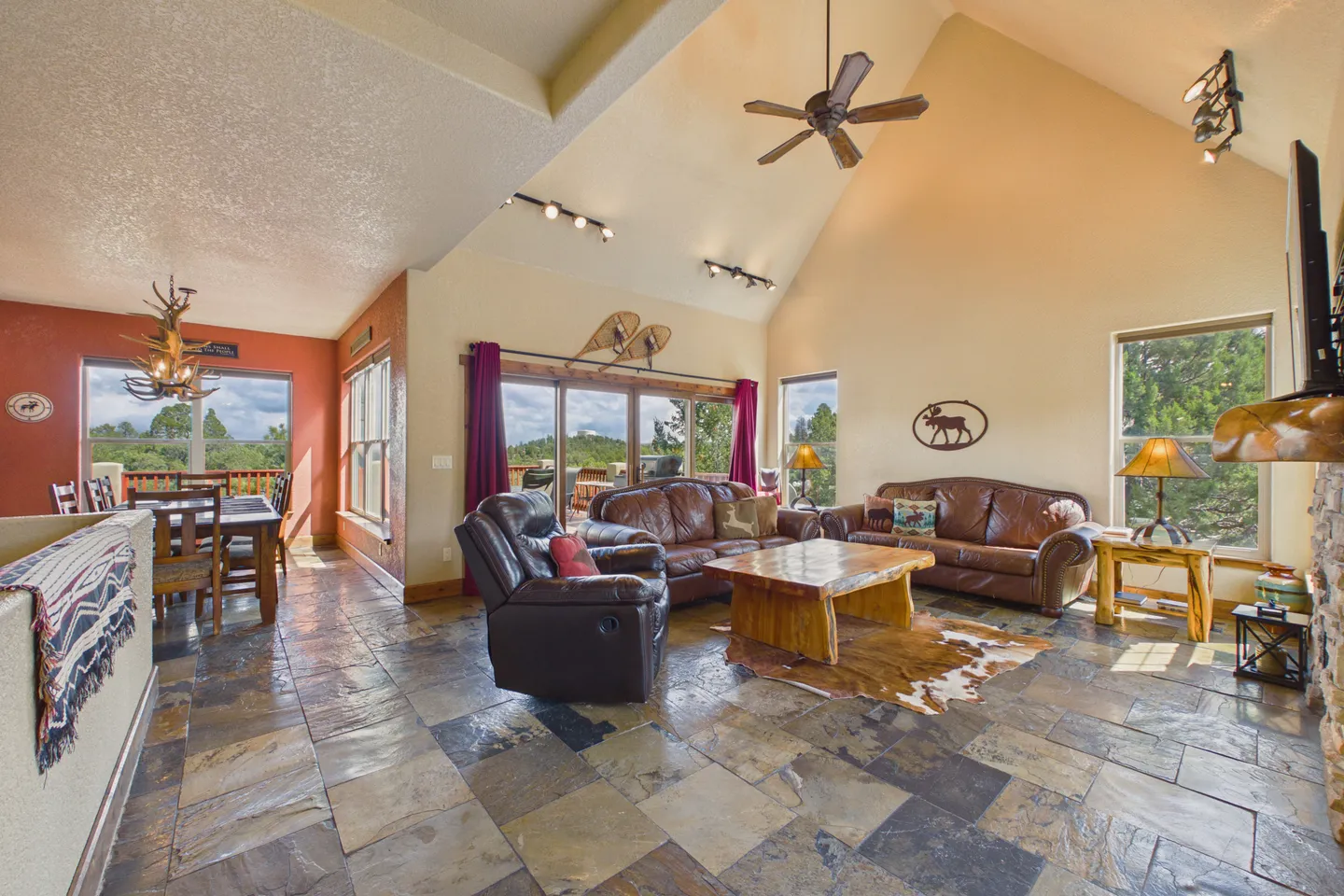 Open-concept living room and dining area with high vaulted ceilings, tiled flooring, leather seating, and sliding glass doors leading to an outdoor deck.