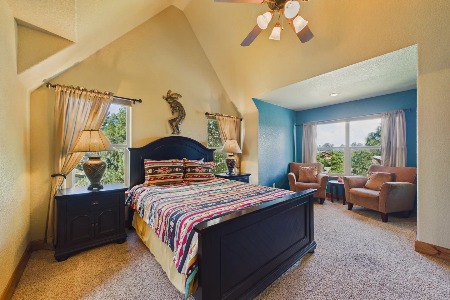 Bedroom with vaulted ceiling, patterned bedding, dark wood bed and nightstands, and a sitting area with two armchairs.