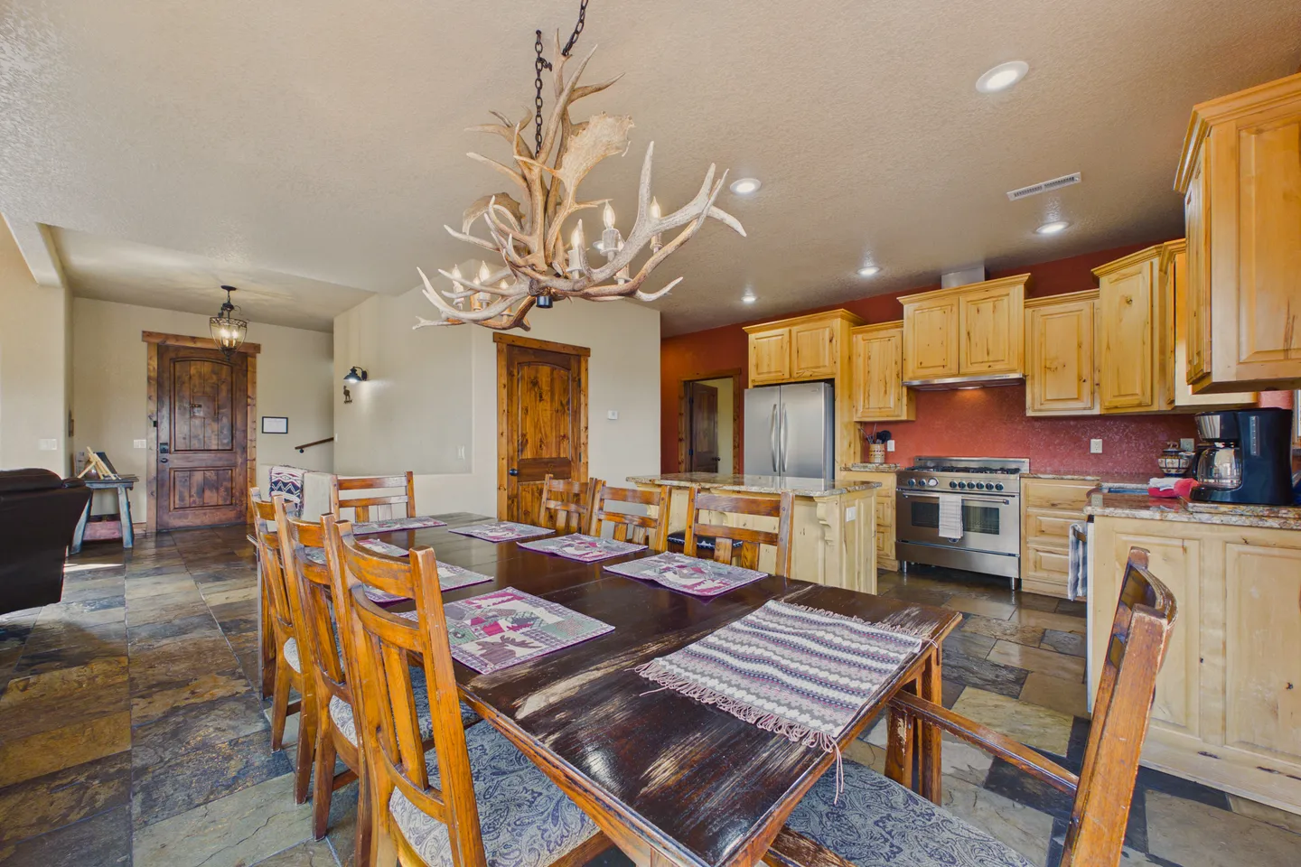 Dining area with a large wooden table and antler chandelier, adjacent to an open-concept kitchen with light wood cabinets.