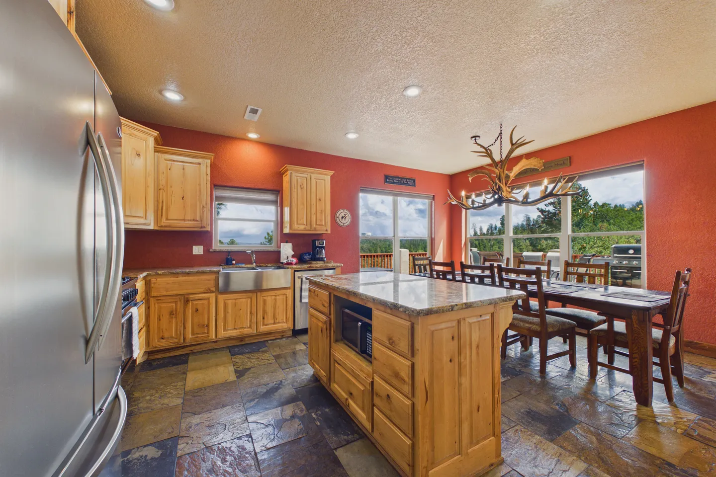 Open-concept kitchen and dining area featuring natural wood cabinetry, a center island, slate tile flooring, red walls, and an antler chandelier over the dining table.