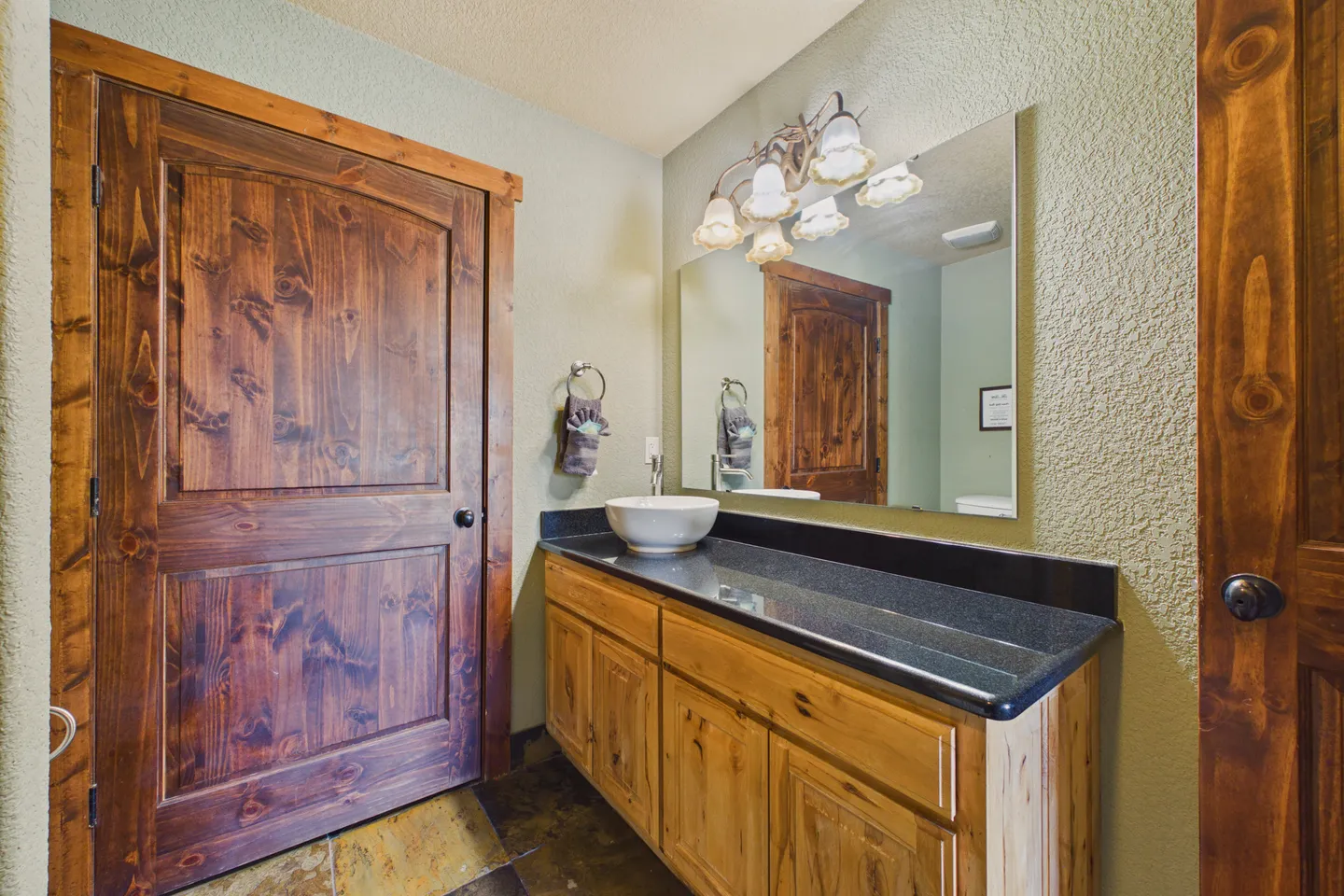 Bathroom with a dark countertop vanity, light wood cabinets, vessel sink, large mirror, and rustic dark wood door.