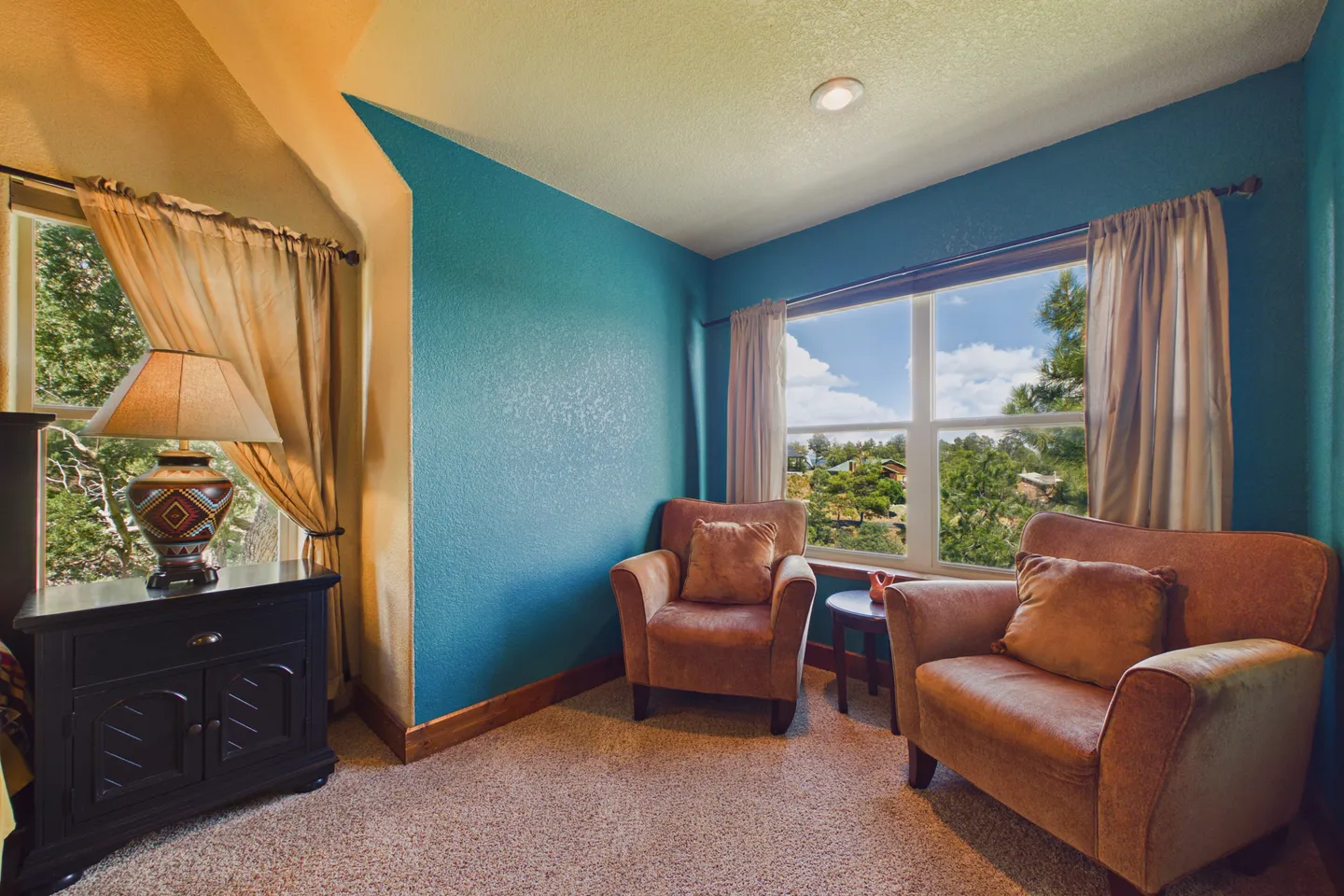 Bedroom sitting area with teal blue accent walls, carpeted flooring, two armchairs near a large window, and a dark nightstand with a lamp.