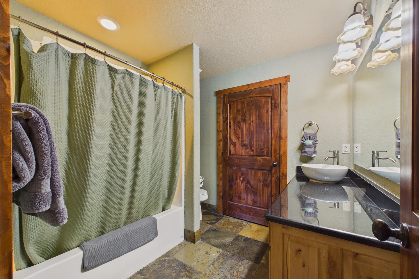 Bathroom featuring a tub-shower combination with a green curtain, a slate tile floor, a rustic wood door, and a vanity with a vessel sink.
