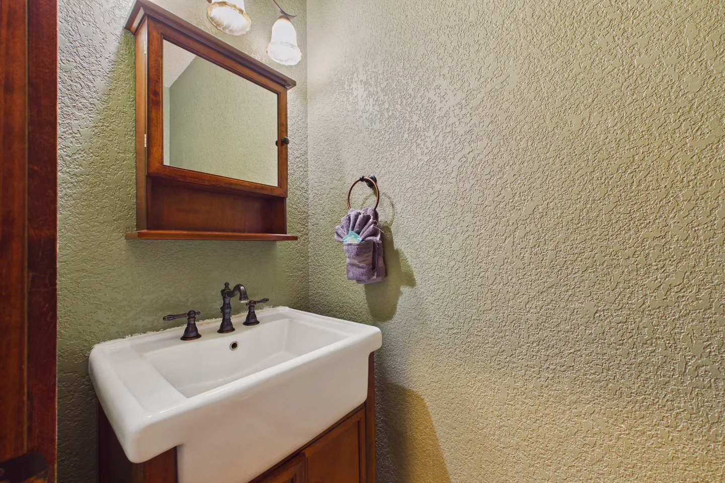 A bathroom vanity with a white ceramic sink, dark faucet, wooden cabinet base, and a matching wooden medicine cabinet on textured green walls.