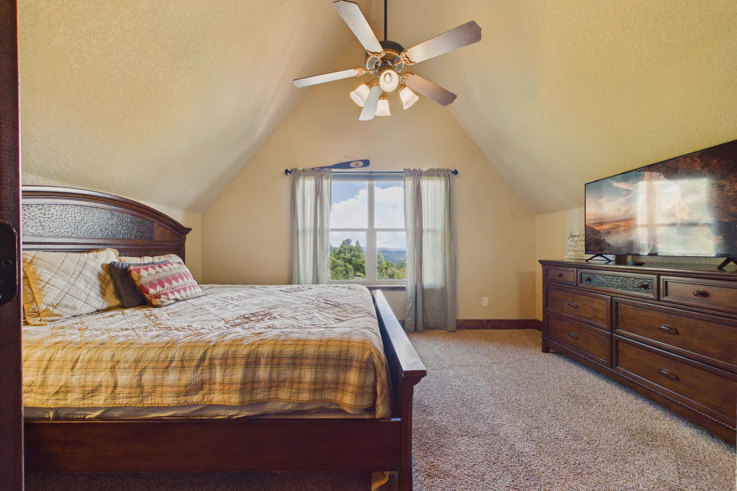 Bedroom with vaulted ceiling, wooden bed, dresser with TV, carpeted floor, and a window with mountain views.