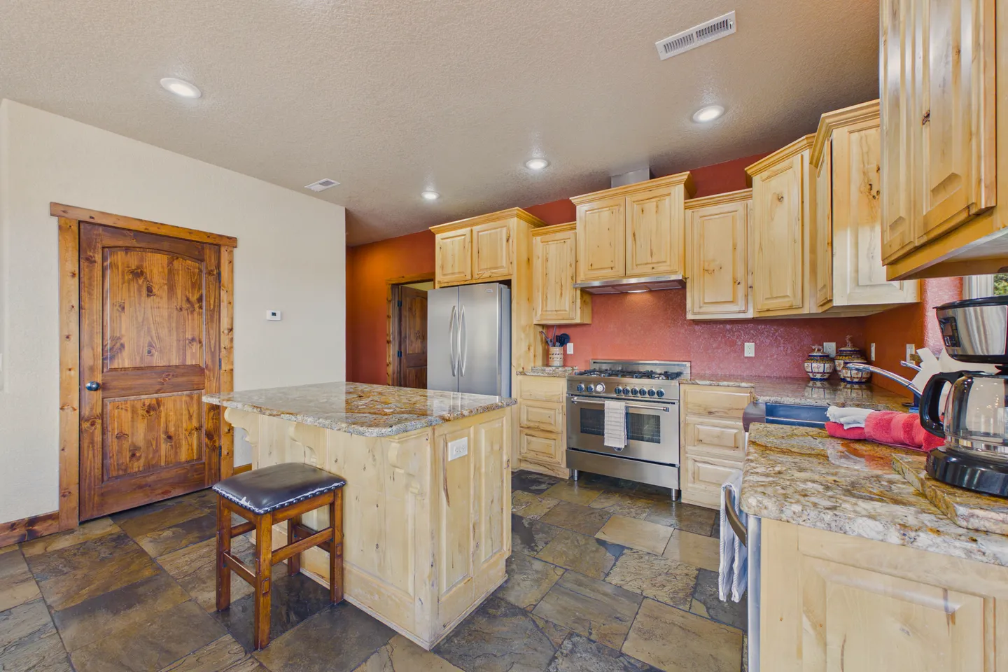 Kitchen with light wood cabinetry, stone countertops, stainless steel appliances, a kitchen island with a stool, and tiled floors.