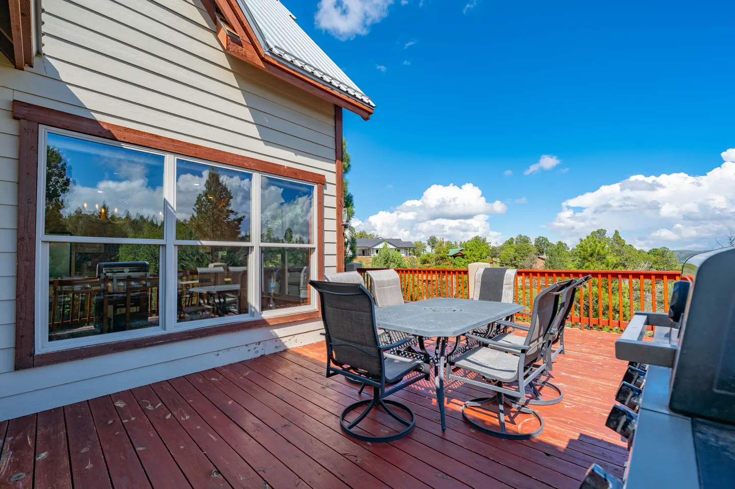 Outdoor wooden deck with a patio dining table, swivel chairs, a barbecue grill, and views of trees under a blue sky.