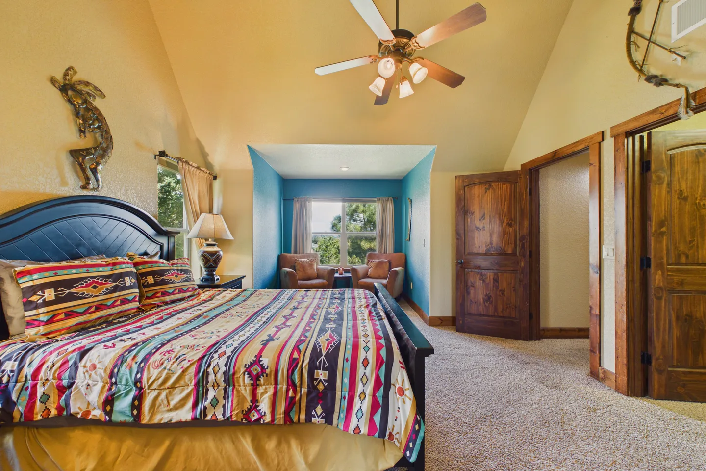 Bedroom with vaulted ceiling, patterned bedding, dark wood bed frame, ceiling fan, and an alcove seating area with two armchairs.