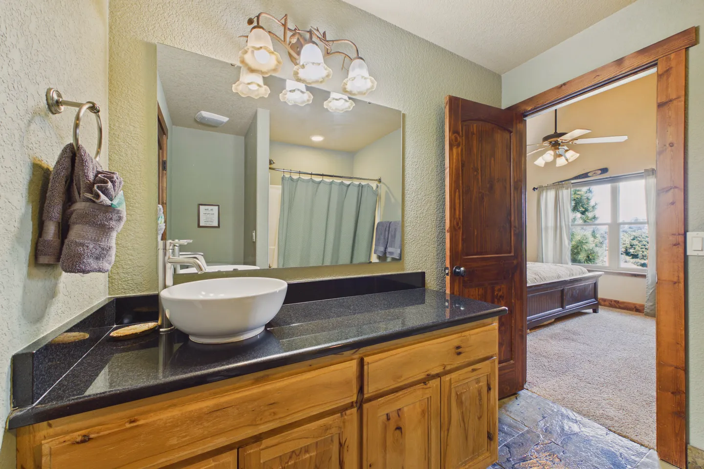 Bathroom with a dark countertop, white vessel sink, wooden vanity, and an open doorway looking into a bedroom.