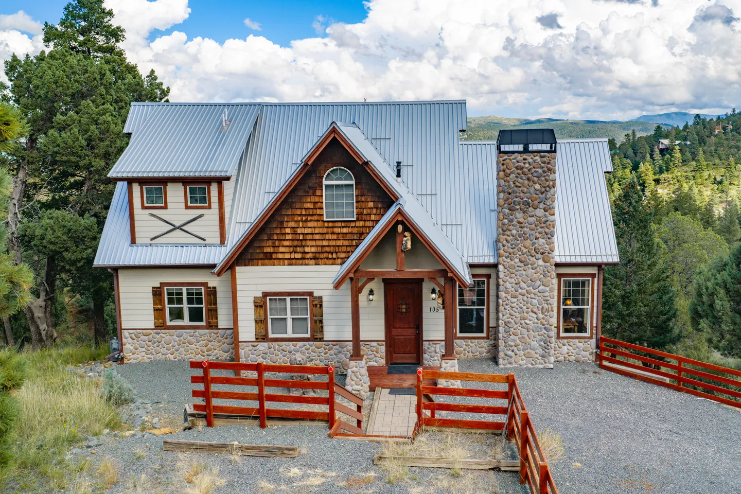 Exterior front view of a two-story home with a metal roof, river stone chimney, wood shake siding details, and a red wooden fence.