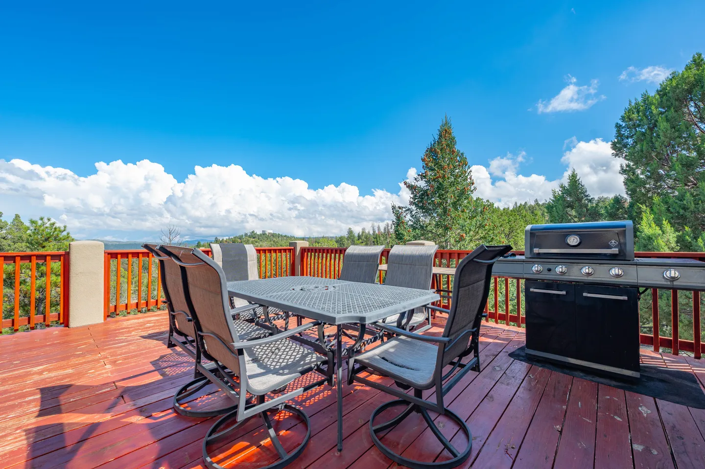 Outdoor wooden deck featuring a dining table with chairs, a gas grill, and views of surrounding pine trees and sky.