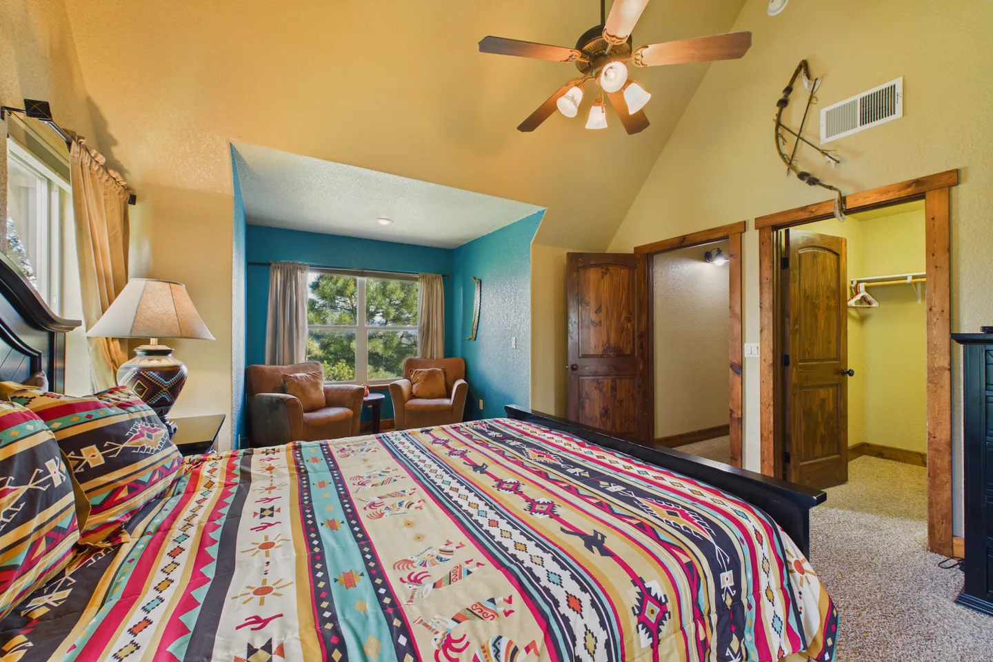 Bedroom with a patterned bedspread, vaulted ceilings, ceiling fan, and an alcove seating area with two armchairs.