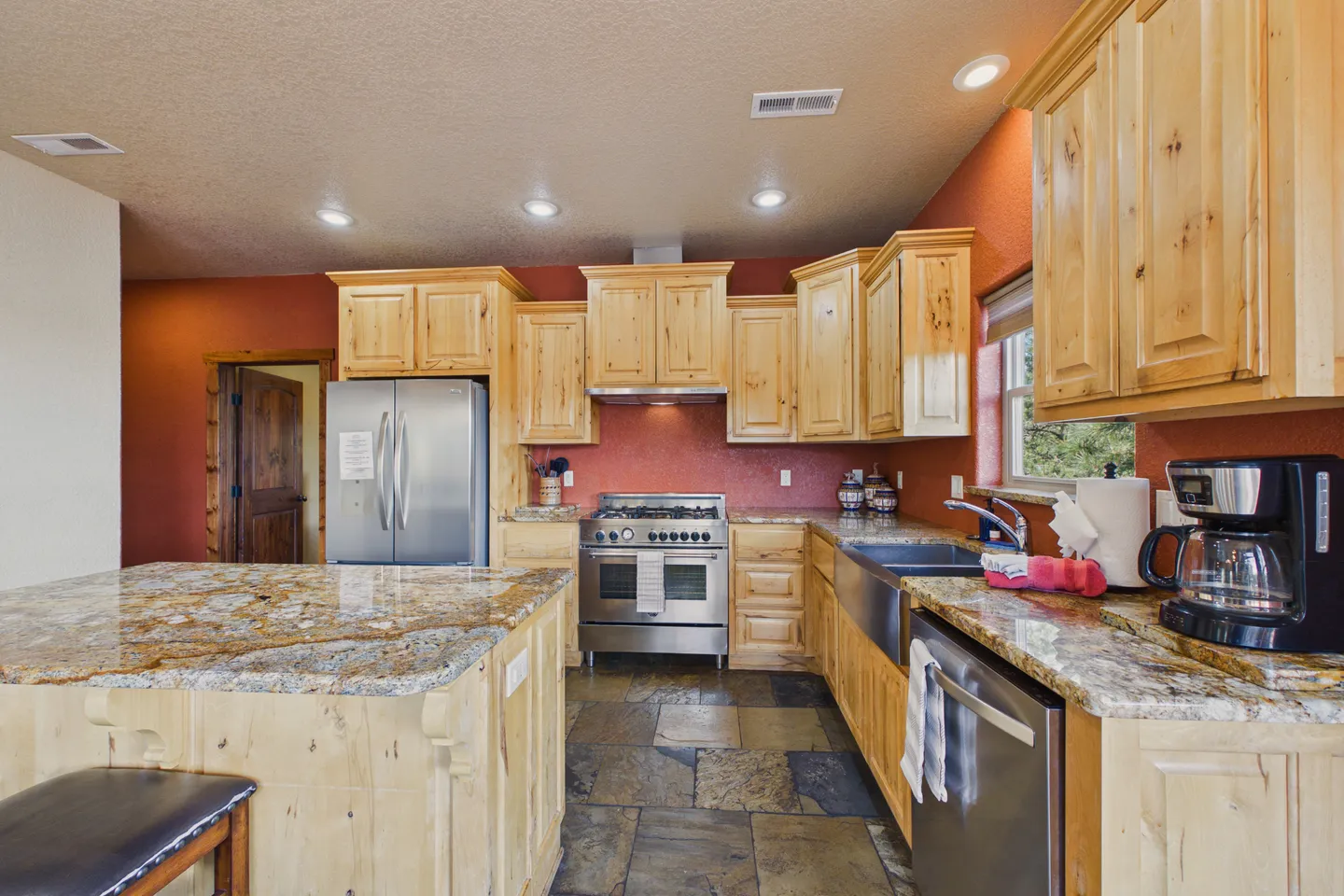 Kitchen with light wood knotty cabinets, granite countertops, stainless steel appliances, and slate tile floors against terracotta-colored walls.