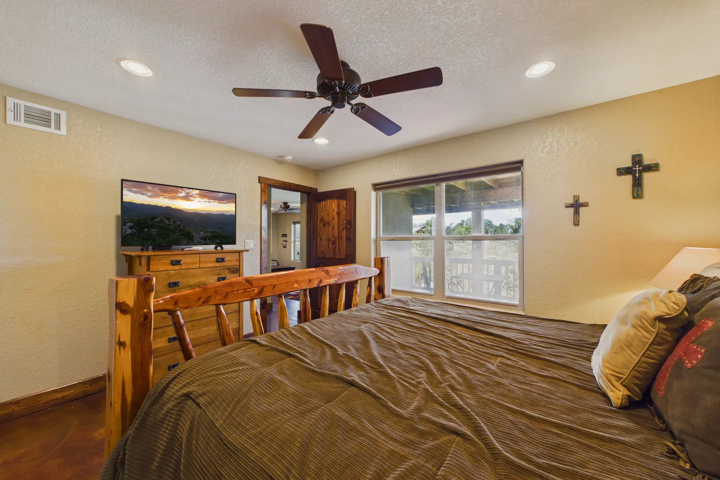 Bedroom featuring a wooden log-style bed, a ceiling fan, a dresser with a wall-mounted TV, and a window overlooking a deck.