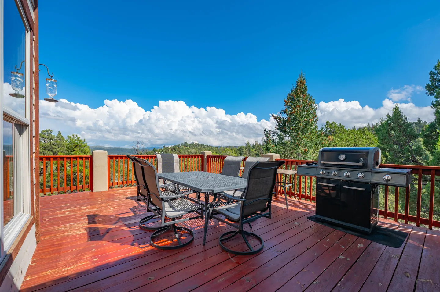 Spacious wooden outdoor deck with a patio dining set, barbecue grill, and panoramic mountain views under a bright blue sky.