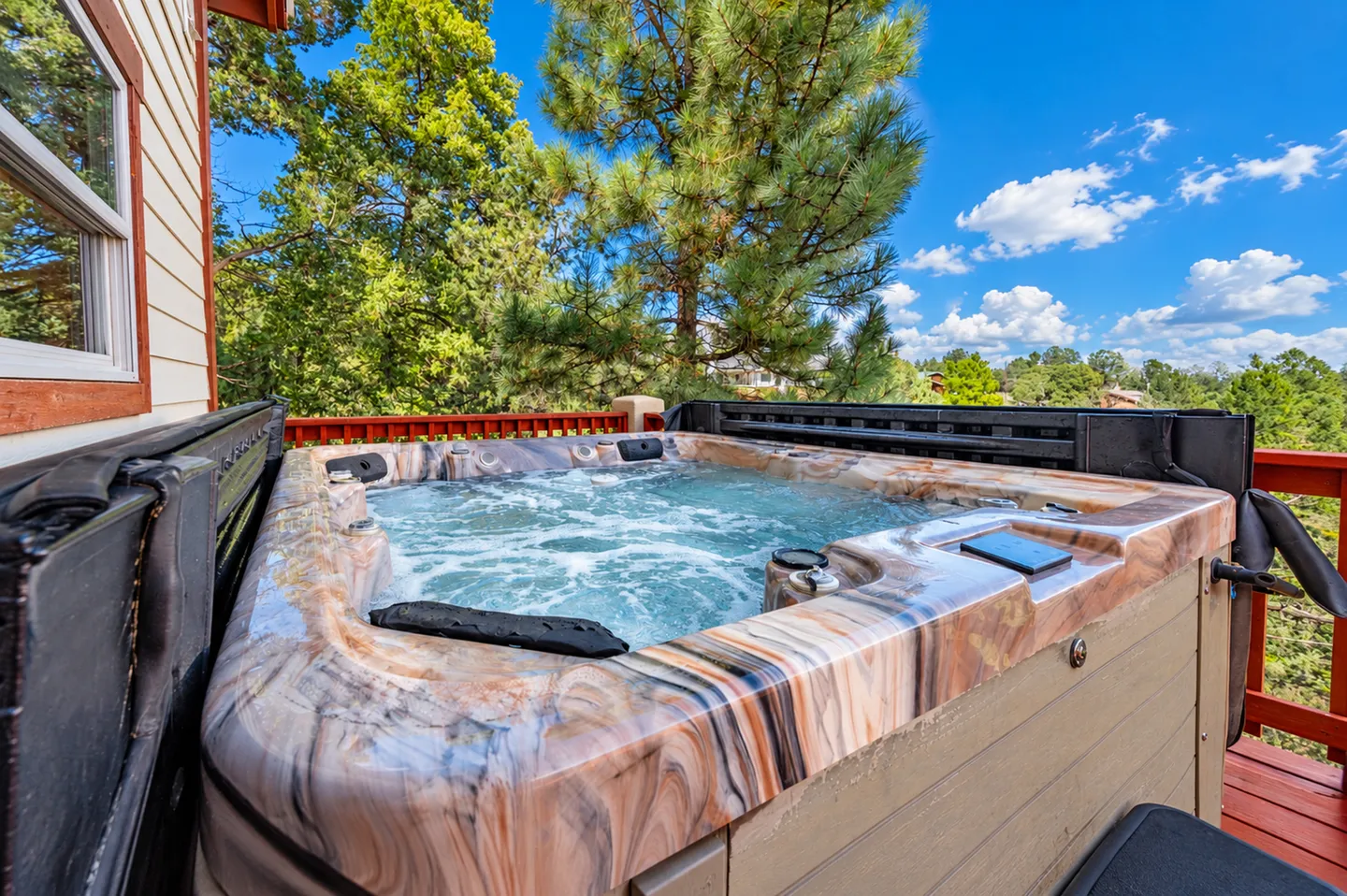 Outdoor hot tub filled with bubbling water on a deck surrounded by pine trees under a blue sky.