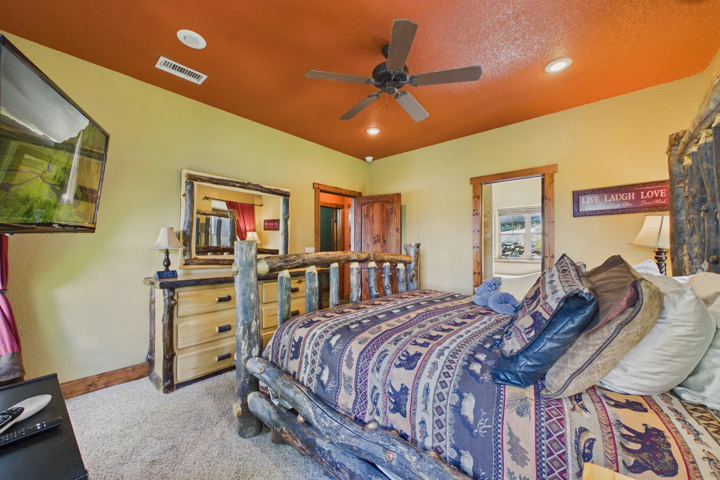 Rustic bedroom with a log-frame bed, matching log-framed dresser with mirror, ceiling fan, and wall-mounted television.