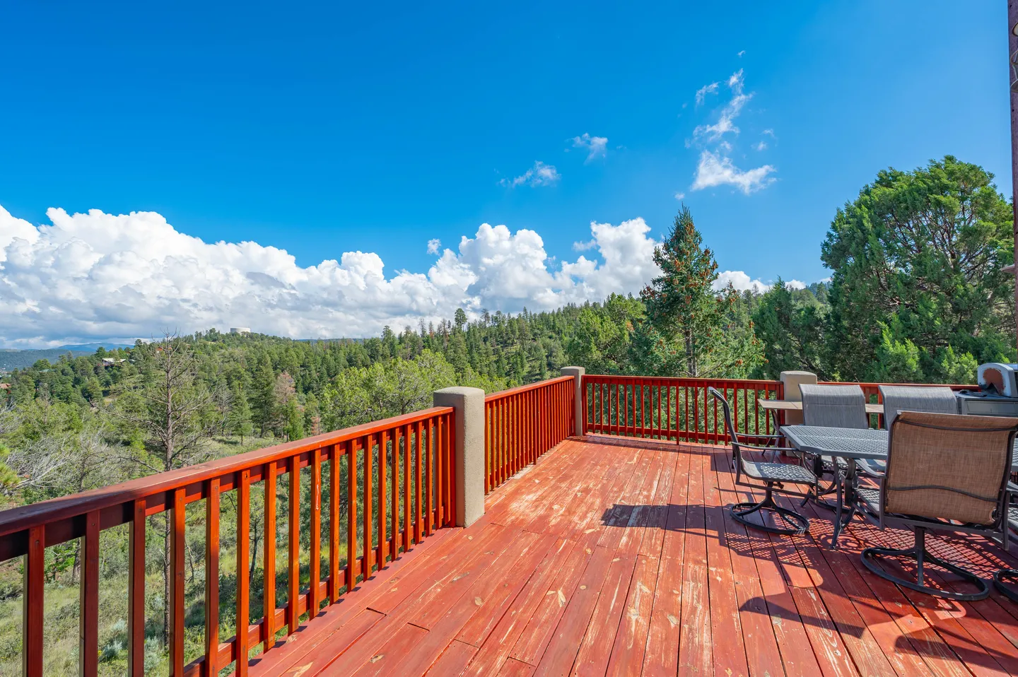 Elevated wooden deck with outdoor dining table and chairs overlooking forested hills under a blue sky.