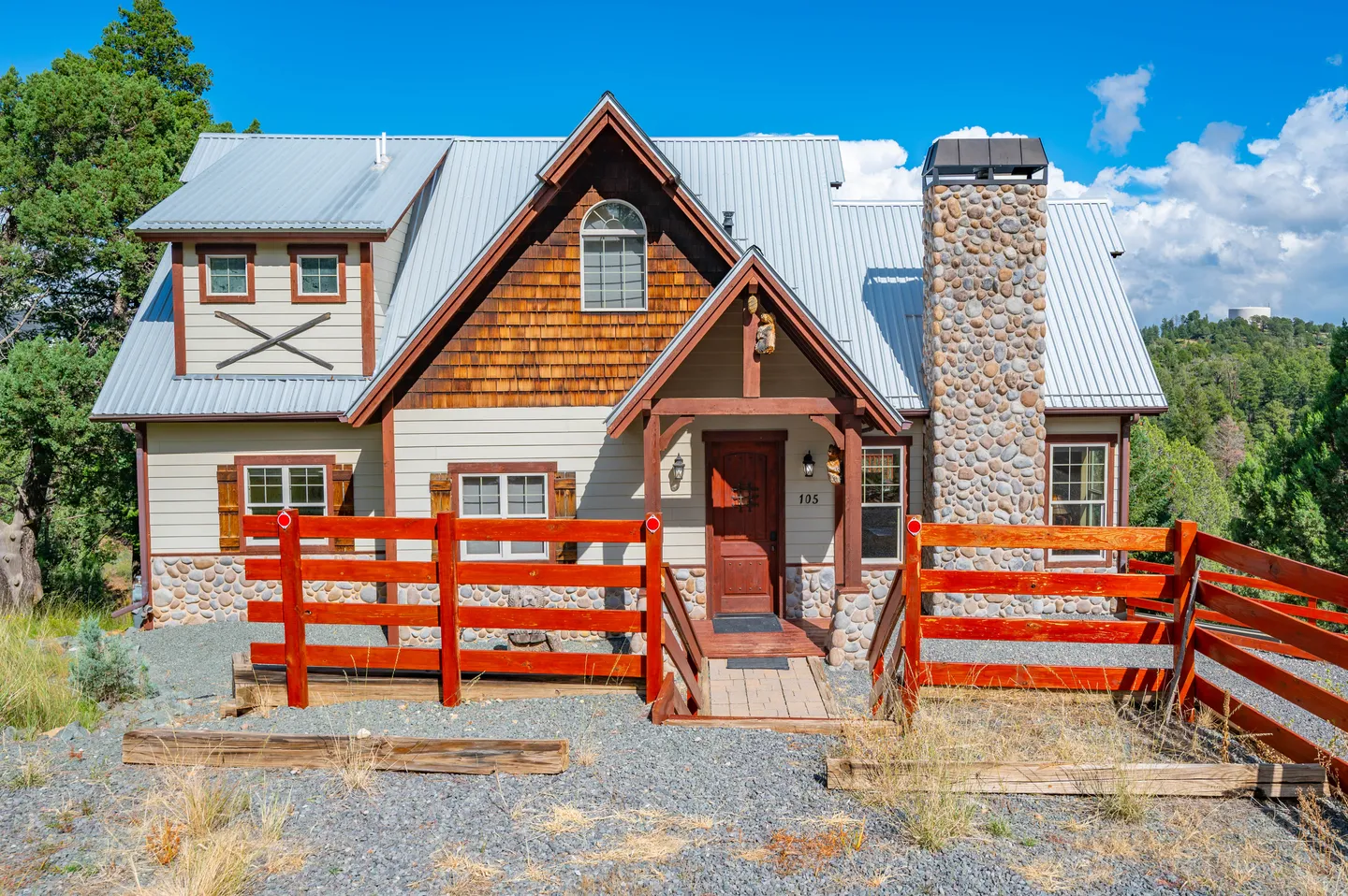 Exterior view of a multi-story cabin-style home with a metal roof, stone chimney, wood siding, and reddish-orange fencing.