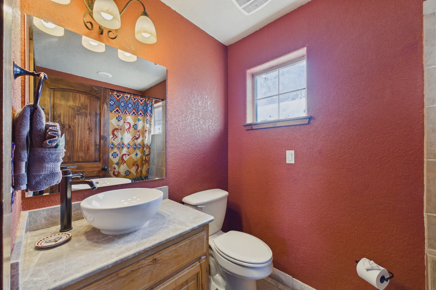 Bathroom featuring warm rust-red walls, a wooden vanity with a white vessel sink, a toilet, and a small high window.