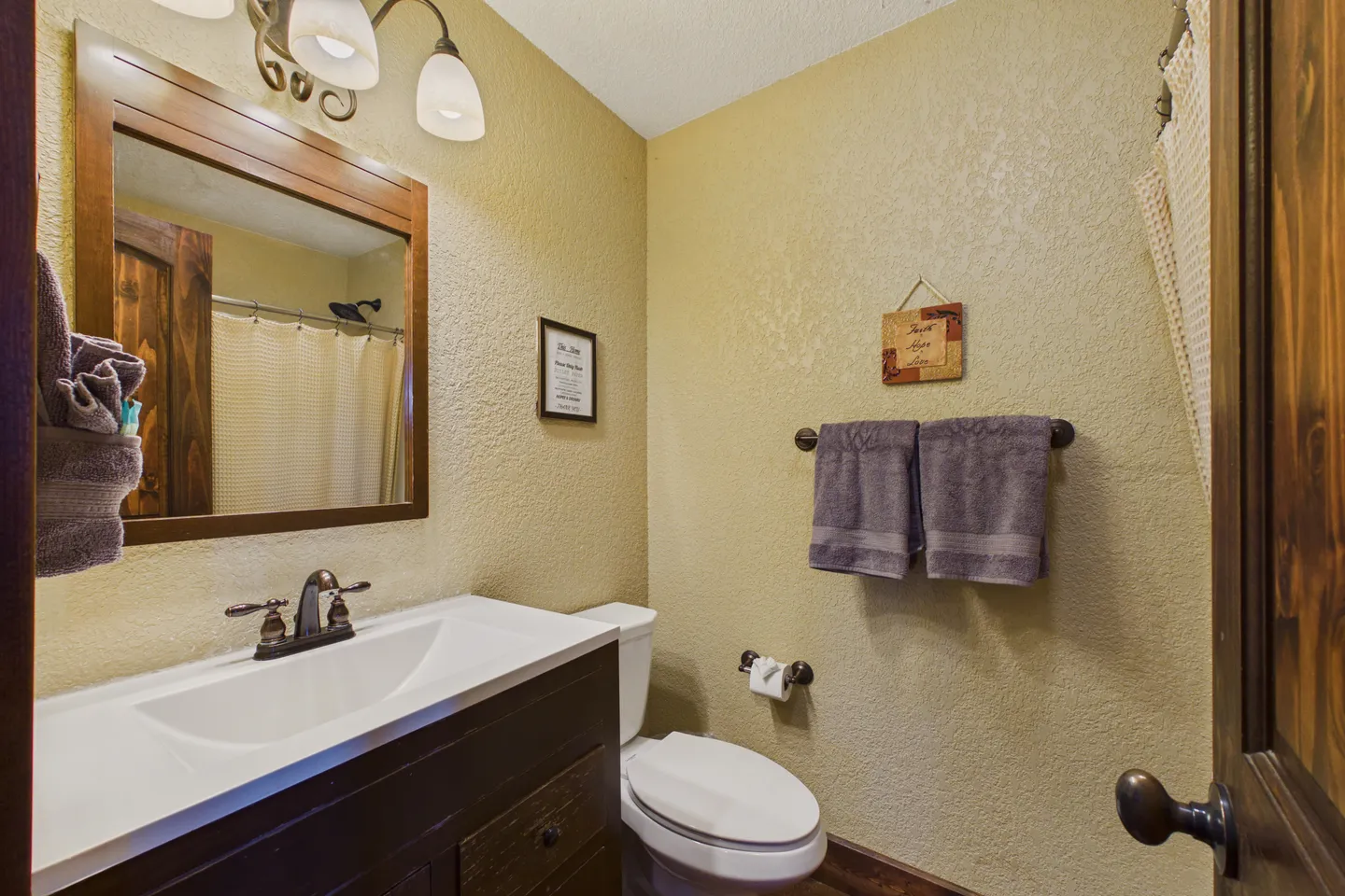 Bathroom featuring a dark wood vanity with a white countertop, framed mirror, toilet, towel bar with grey towels, and light-colored textured walls.