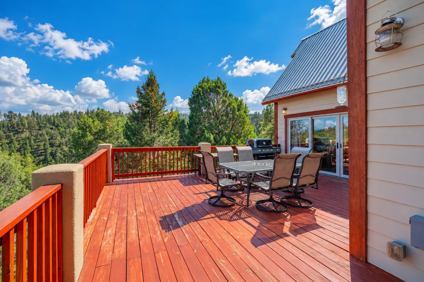 Sunny wooden deck with dining patio table, chairs, barbecue grill, and view of evergreen trees under a bright blue sky.