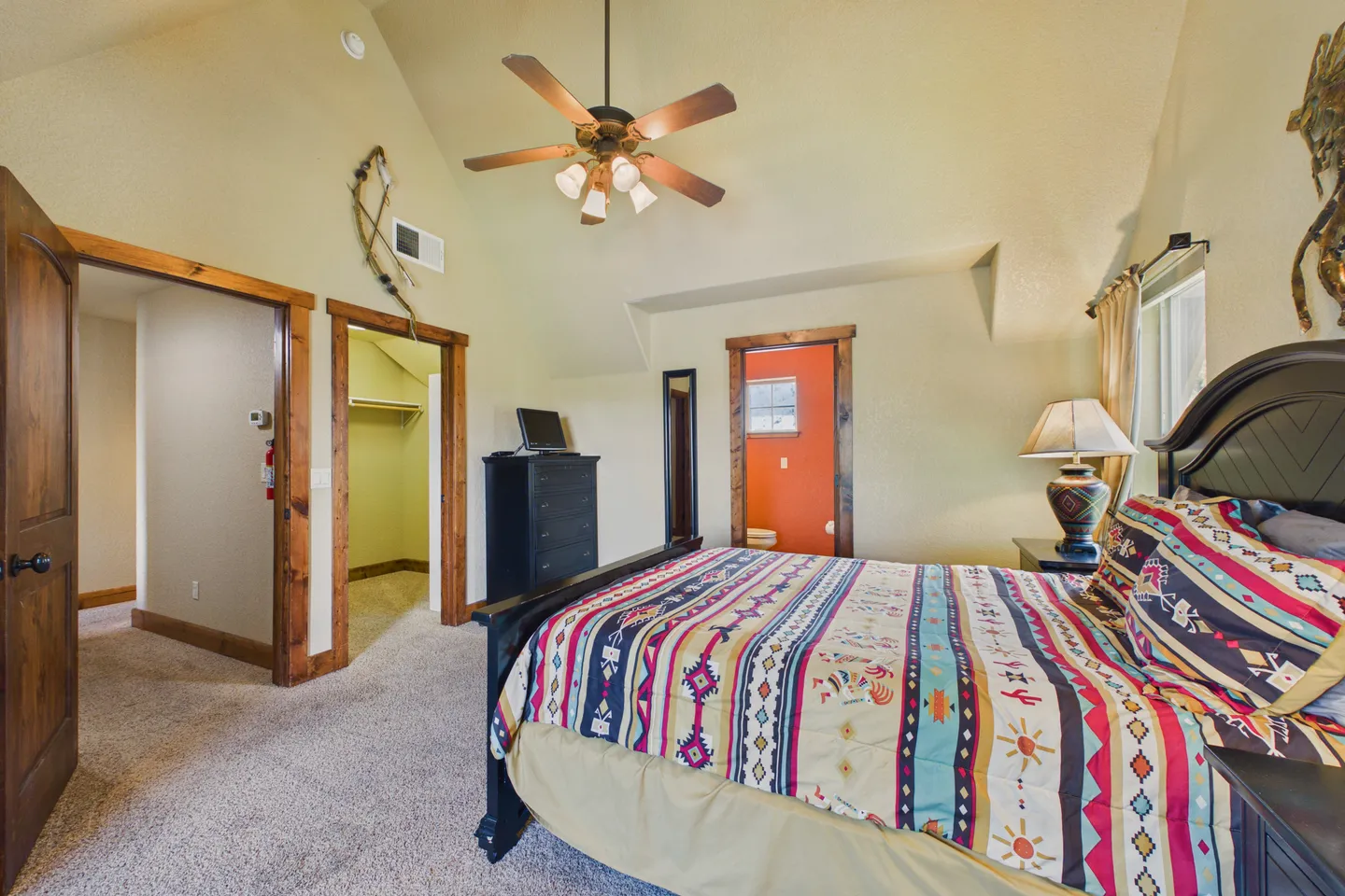Bedroom with vaulted ceiling, ceiling fan, carpeted floor, patterned bedding, and doorways to a hallway, closet, and bathroom.