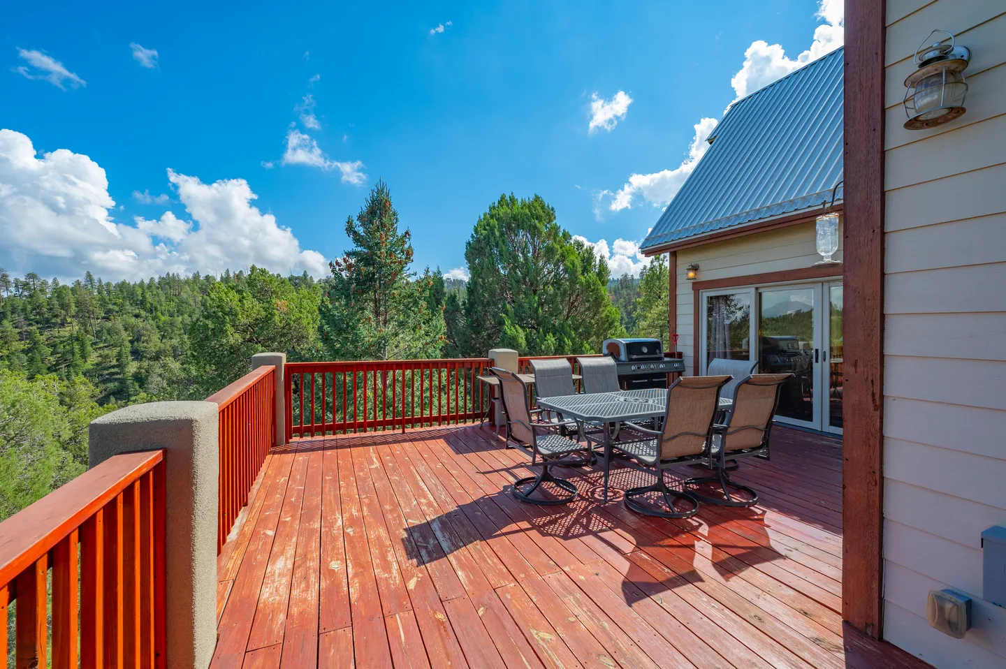 Sunlit wooden deck with an outdoor dining table, chairs, and barbecue grill overlooking a pine forest under a blue sky.